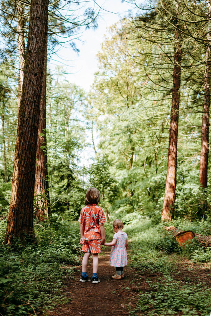 Bruder steht mit seiner kleinen Schwester an der Hand auf dem dichtbewachsenen Waldweg.