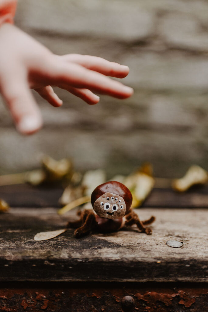 Eine Kinderhand streckt sich nach einer selbstgebastelten Kastanien-Spinne aus, die auf einer Holzbank steht und eine herbstliche Bastelszene darstellt.