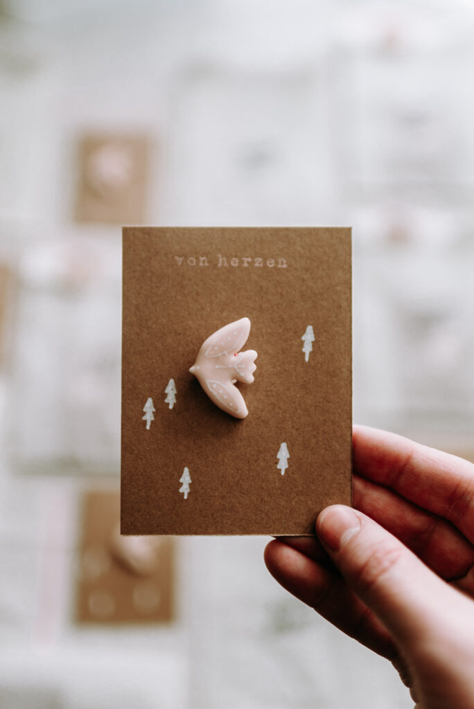 Detailaufnahme eines roséfarbenen Vogel-Ansteckers aus Clay (Polymer Clay) auf einer braunen Karte mit der Aufschrift „von herzen“.