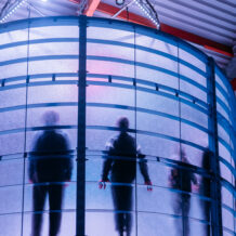 Mehrere Personen als dunkle Silhouetten hinter einer blauen Glasfassade.