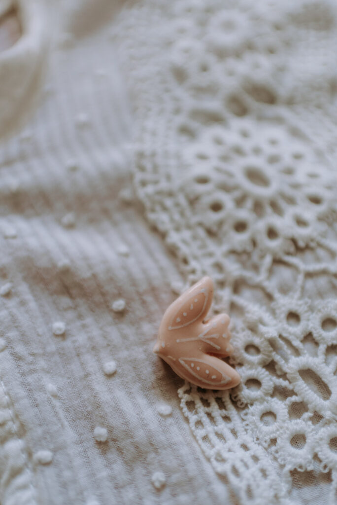 Roséfarbener Vogel-Anstecker aus Clay (Polymer Clay) liegt auf hellem Stoff mit feiner Spitzenstruktur.