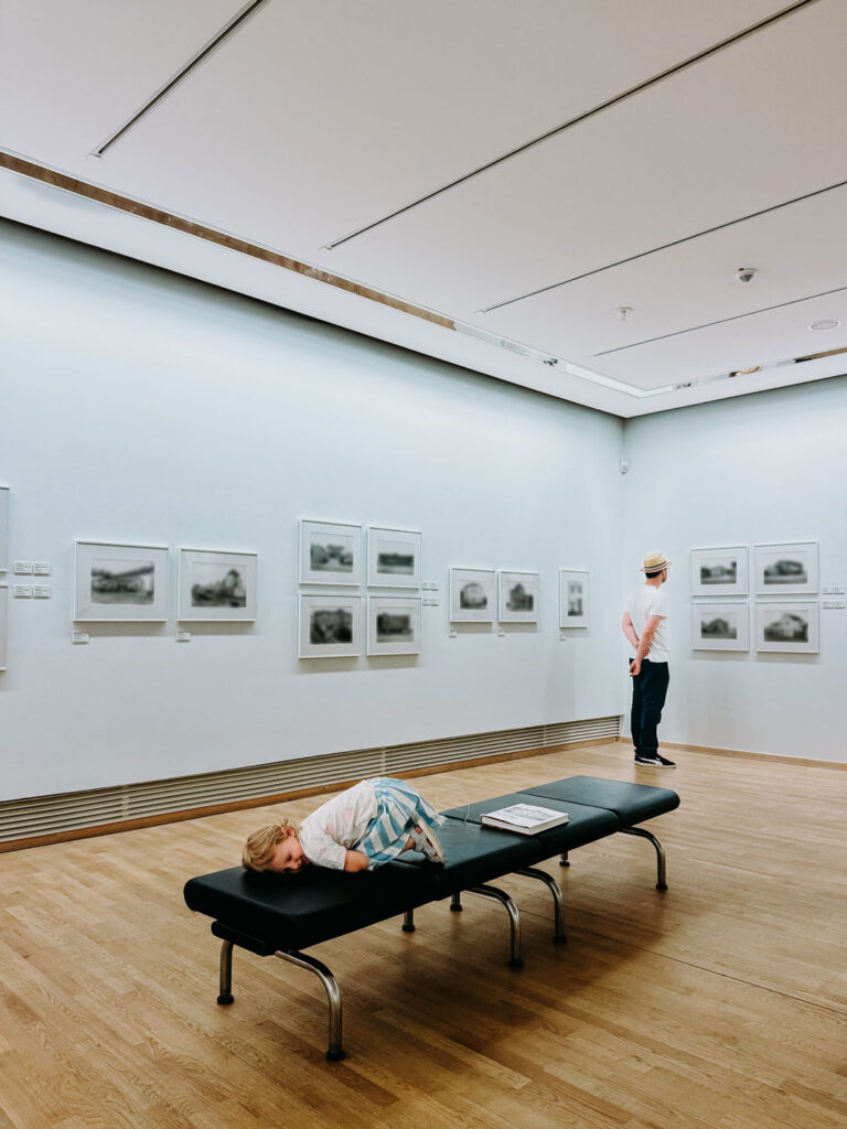 Kind liegt auf einer Bank in einem Museumsraum, im Hintergrund steht eine erwachsene Person