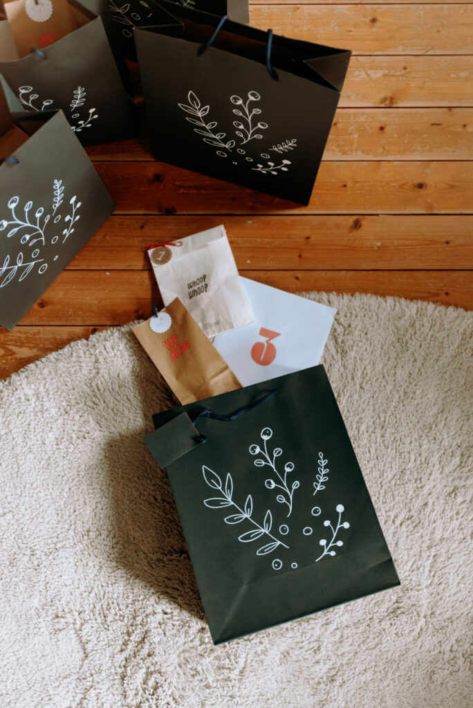 Mehrere schwarze Papiertüten mit weißen floralen Doodle-Motiven stehen auf einem Holzboden. Davor liegen kleine Papiertütchen als Adventsgeschenke.