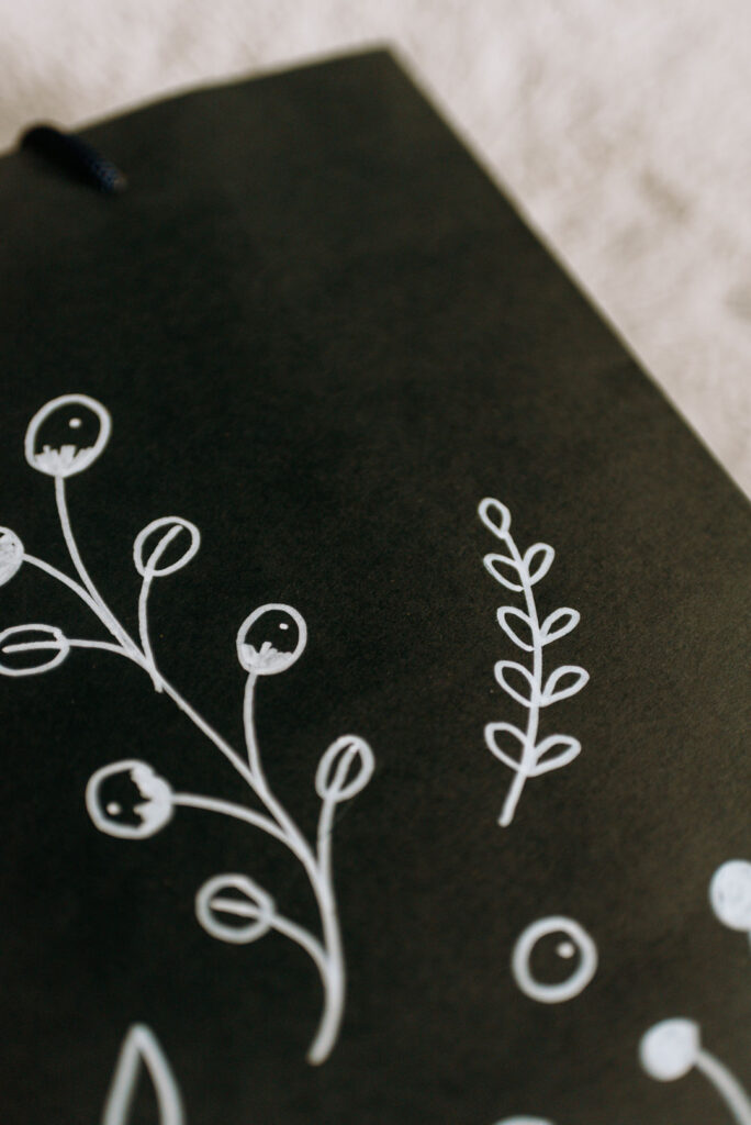 Nahaufnahme eines handgezeichneten weißen floralen Doodle-Zweigs auf einer schwarzen Papiertüte.
