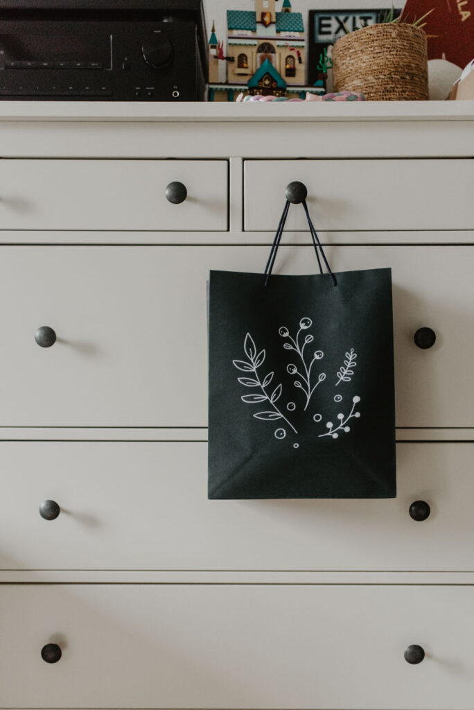 Eine schwarze Geschenktüte mit weißem floralen Doodle-Motiv hängt an einer hellen Kommode.
