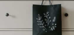Eine schwarze Geschenktüte mit weißem floralen Doodle-Motiv hängt an einer hellen Kommode.